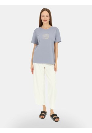 Kobieta stojąca przodem, ubrana w jasnoniebieski t-shirt z kwadratowym wzorem, białe krótkie spodnie i czarne sandały na jednolitym tle.