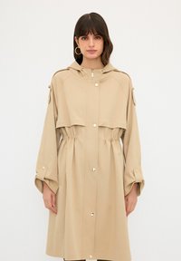 Beige regnkappa med huva, elastisk midja och tryckknappsstängning. Har vida ärmar och en kortad överdeldesign. Slät textur.