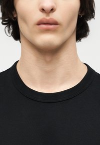 Gros plan sur la partie inférieure du visage et le cou d'une personne portant un t-shirt noir à col rond, avec de courts cheveux foncés et des boucles d'oreilles cerclées.