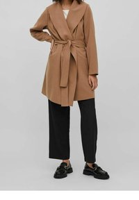 Manteau marron style portefeuille avec ceinture nouée, manches longues et poches latérales, associé à un pantalon noir à jambes larges et des mocassins noirs.