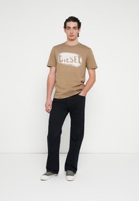 Tan katoenen T-shirt met een spuitverf "DIESEL" logo, gecombineerd met donkerblauwe broek en grijze sneakers met witte zolen. Eenvoudig ontwerp.