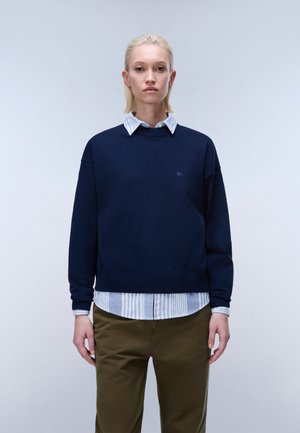 Junge Person mit kurzen blonden Haaren, trägt einen marineblauen Pullover über einem gestreiften Hemd und olivgrüne Hose vor einem schlichten grauen Hintergrund.