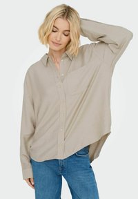 Beige košile s dlouhým rukávem na knoflíky, mírně oversized, s kapsou na prsou, vyrobená z lehkého materiálu, kombinovaná s modrými džíny.