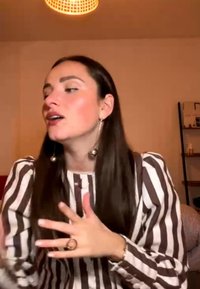 Donna con lunghi capelli scuri e camicia a righe che gesticola con le mani mentre parla all'interno sotto una luce sospesa.