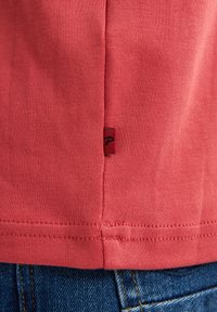 Koraalkleurig katoen T-shirt met een zijnaadlabel en een klein rood embleem, gecombineerd met blauwe denim jeans. Vlotte textuur en eenvoudig ontwerp.