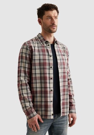 Mann trägt ein rot-weiß-schwarz kariertes Hemd mit langen Ärmeln über einem schwarzen T-Shirt und blauen Jeans, steht vor einem schlichten hellen Hintergrund.