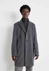 Cappotto in misto lana grigio con colletto a risvolto, chiusura con un solo bottone e tasche laterali, indossato sopra una giacca nera con zip.