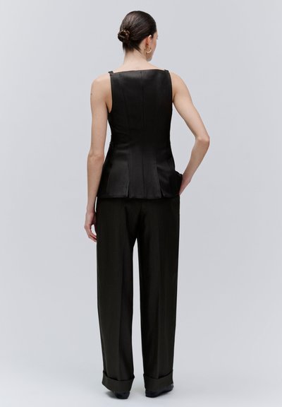 Vrouw met haar in een knot draagt een mouwloze, nauwsluitende zwarte top en zwarte broek met wijde pijpen, staande met haar rug naar de camera tegen een effen achtergrond.