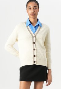 Cream textured cardigan with a V-neckline, brown button closure, and beige ribbed accents. Model wearing a blue shirt and black skirt.