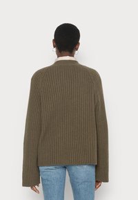 Persona con capelli corti e orecchini a cerchio, che indossa un maglione a coste verde oliva e jeans azzurri chiari, di spalle su uno sfondo semplice.