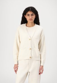 Strikket creme cardigan med lange ærmer, v-hals, to forkotalet lommer og guldknapper. Båret over en lys top og parret med beige bukser.