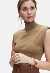 Bruine geribde mouwloze top met hoge kraag, gecombineerd met een bruine broek. Accessoires omvatten gouden oorbellen en een horloge met een leren band.