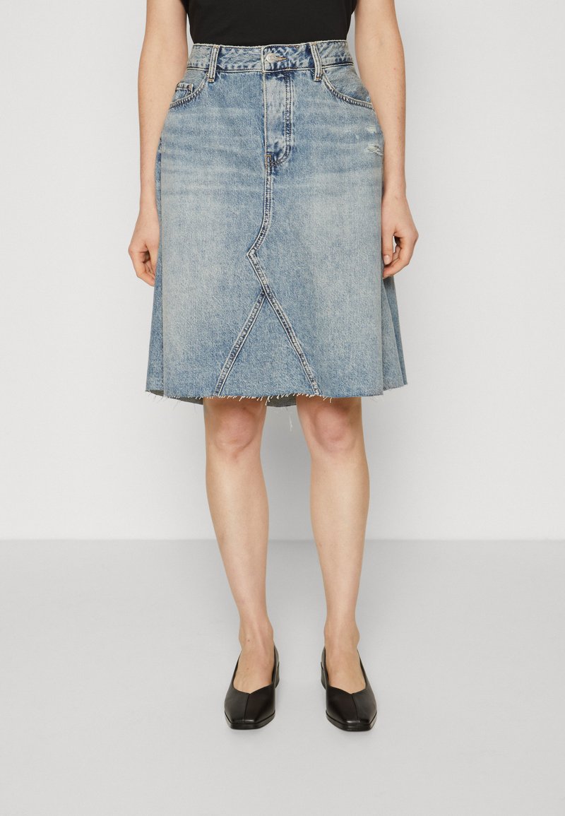 Jeanskjørt i lys blå, til knærne, med frynset kant og skrå sømdetaljer. Brukt med svarte slip-on sko.
