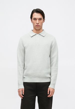 Pull en gris clair avec un tissage texturé, présentant un col zippé et un col plat. Les poignets et l'ourlet sont côtelés. Porté avec un pantalon sombre.