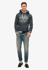 Felpa con cappuccio blu scuro con una grafica di una montagna e lacci bianchi, abbinata a jeans azzurri, scarpe nere e un orologio.