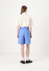 Svetlá béžová košeľa s krátkym rukávom a modré strihané šortky s gombíkovými akcentmi. Čierne topánky a biele členkové ponožky dokončujú outfit.