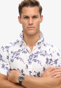 Camisa de manga corta en blanco con un patrón floral púrpura, con cuello abotonado. En la muñeca hay un reloj oscuro con correa de cuero.