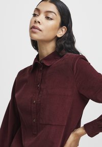 Burgundi värvi vampiirkangas särk nööbiga kaelusega, rinnaosal tasku ja pikad varrukad. Omab pruune nööpe ja sujuvat tekstuuri.