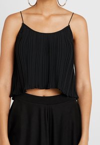 Femme portant un haut court à fines bretelles plissé noir et un pantalon noir taille haute, avec de grandes boucles d'oreilles créoles visibles.