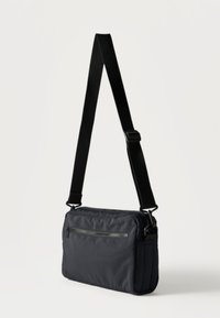 Μικρό μαύρο τσαντάκι ώμου με ρυθμιζόμενο ιμάντα και εμπρόσθια τσέπη με φερμουάρ, τοποθετημένο σε λευκό φόντο.