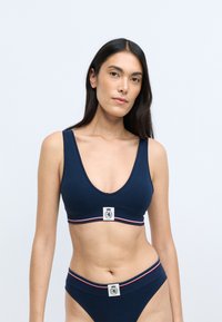Navy blue ribbed bralette with a deep V-neck and wide straps, featuring a logo patch and contrasting red and white stripes on the band.