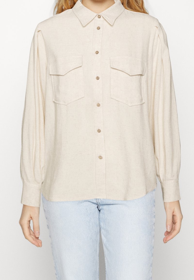 Femme portant une chemise crème boutonnée avec deux poches à rabat sur la poitrine et des manches longues plissées, associée à un jean bleu clair.
