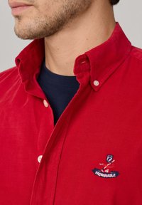 Rode corduroy overhemd met button-down kraag. Kenmerkt een geborduurd logo en een marineblauwe binnenlaag zichtbaar onder de kraag.