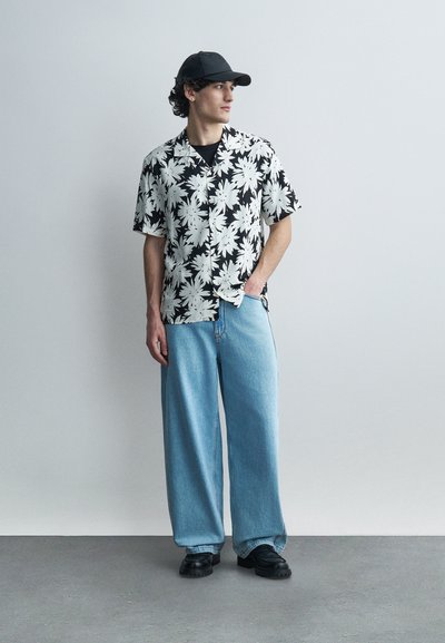 Jeune homme portant une casquette noire, une chemise à manches courtes florale noir et blanc, un jean large bleu clair et des chaussures noires, debout contre un mur uni.