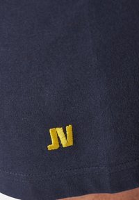 Navy cotton fabric with a smooth texture featuring a yellow embroidered "JN" logo near the hem. Simple, clean design focused on the logo.