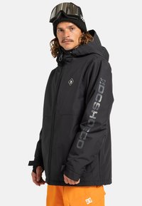 Muž v čiernej snowboardovej bunde značky DC s kapucňou, čiernej čapici a okuliaroch, a jasne oranžových snowboardových nohaviciach značky DC, hľadiaci dopredu s neutrálnym výrazom.