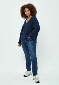 Navy-blauer, gerippter Strickpullover mit V-Ausschnitt, kombiniert mit blauen Jeans und grauen Turnschuhen. Das Outfit zeichnet sich durch eine lockere Passform und strukturierter Stoff aus.