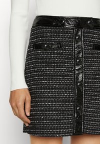 Zwart-wit tweed rok met een glanzende zwarte tailleband, voorzien van voordeeltjes en twee kleine zakken met zwarte knopen.