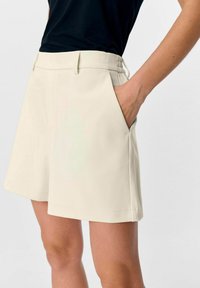 Beige op maat gemaakte shorts met zijzakken, omgeslagen zomen en een gladde stoftextuur. G gedragen met een zwart top.