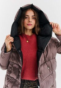 Puszysta, metaliczna mauve kurtka z czarną podszewką i kapturem; posiada pikowany wzór i napy. Nosi się ją na fakturowanym czerwonym swetrze.