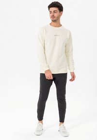 Felpa crema chiara con un piccolo logo nero, abbinata a pantaloni joggers slim-fit grigio scuro con dettagli testurizzati e scarpe da ginnastica bianche.