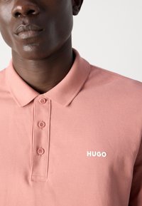 Polo rose en tissu texturé, avec une patte de boutonnage et un logo "HUGO" blanc sur la poitrine. Vue rapprochée de la zone du col.