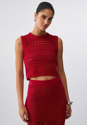 Donna che indossa un crop top all'uncinetto senza maniche rosso e una gonna a vita alta abbinata, accessoriata con orecchini e bracciale d'argento, in piedi davanti a una parete bianca.