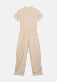 Combinaison beige à manches courtes en tissu doux, dotée d'une taille cintrée, d'un col et de deux poches cargo sur des pantalons à jambes larges.
