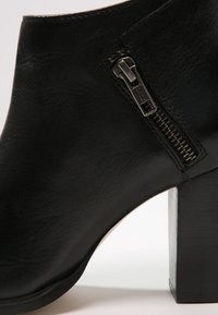 Close-up van een zwarte leren enkelboot met zijketting en stevige blokhak op een witte achtergrond.