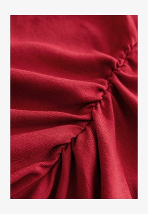 Piros szövet gyűrt textúrát mutat finom átlós vonalakkal. Az anyag puhanak és enyhén rugalmasnak tűnik, kiemelve a dizájn mélységét.