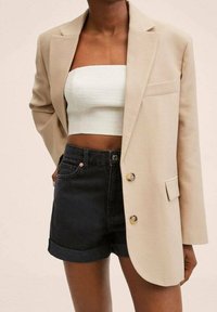 Blazer beige con un solo botón, usado sobre un top blanco ajustado y cropped y shorts de mezclilla negra con dobladillos, con costuras naranjas.