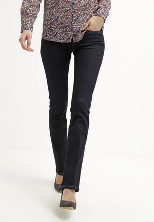 Personne portant un jean slim foncé, une chemise à manches longues à motifs floraux et des chaussures grises à bout pointu, une main dans la poche en marchant.