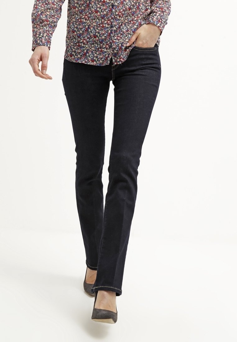 Personne portant un jean slim foncé, une chemise à manches longues à motifs floraux et des chaussures grises à bout pointu, une main dans la poche en marchant.