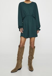 Robe texturée bleu-vert avec de longues manches bouffantes et une taille cintrée, associée à des bottes en daim marron montantes avec des couture décoratives.