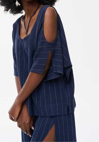 Haut ample bleu marine avec manches découpées, présentant des rayures blanches verticales, un décolleté en V et un tissu doux et texturé.