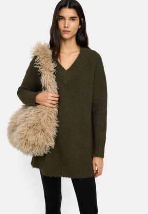 Kobieta ubrana w ciemnozielony, oversize sweter z dekoltem w szpic i czarne spodnie, trzyma dużą beżową puszystą torebkę na białym tle.