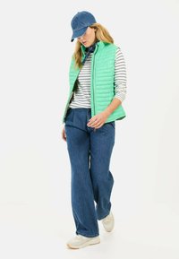Hellgrüne gestepptes Weste über einem weiß-blau gestreiften Oberteil, kombiniert mit weit geschnittenen Jeans und weißen Sneakers, accessoriert mit einer blauen Mütze.