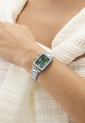 Zilverkleurig metalen horloge met een groen vierkant wijzerplaat en diamanten details, gedragen om de pols tegen een witte, textuurrijke stoffen achtergrond.