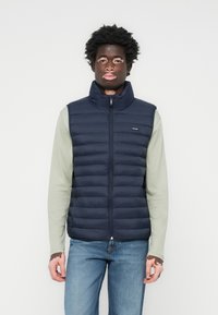 Navy padded vest with high collar and zipper, featuring horizontal quilted design. Worn over a light green long-sleeve shirt and blue jeans.