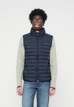 Marine gewatteerd vest met hoge kraag en rits, voorzien van een horizontaal gestikt ontwerp. Draag het over een lichtgroen langemouwen shirt en blauwe spijkerbroek.